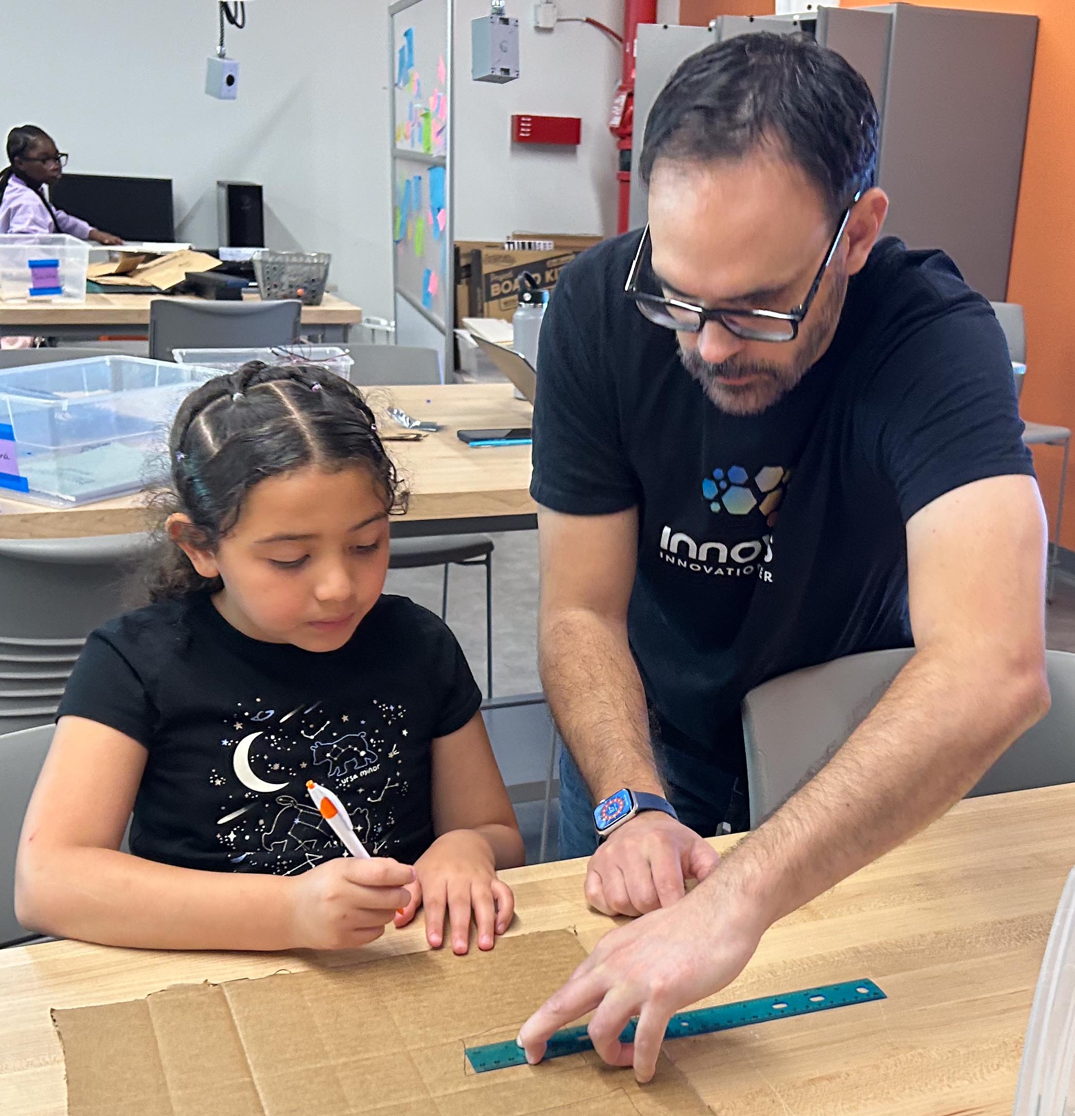 Man and young girl cutting a large cardboard box on a wooden worktable in a bright workshop.